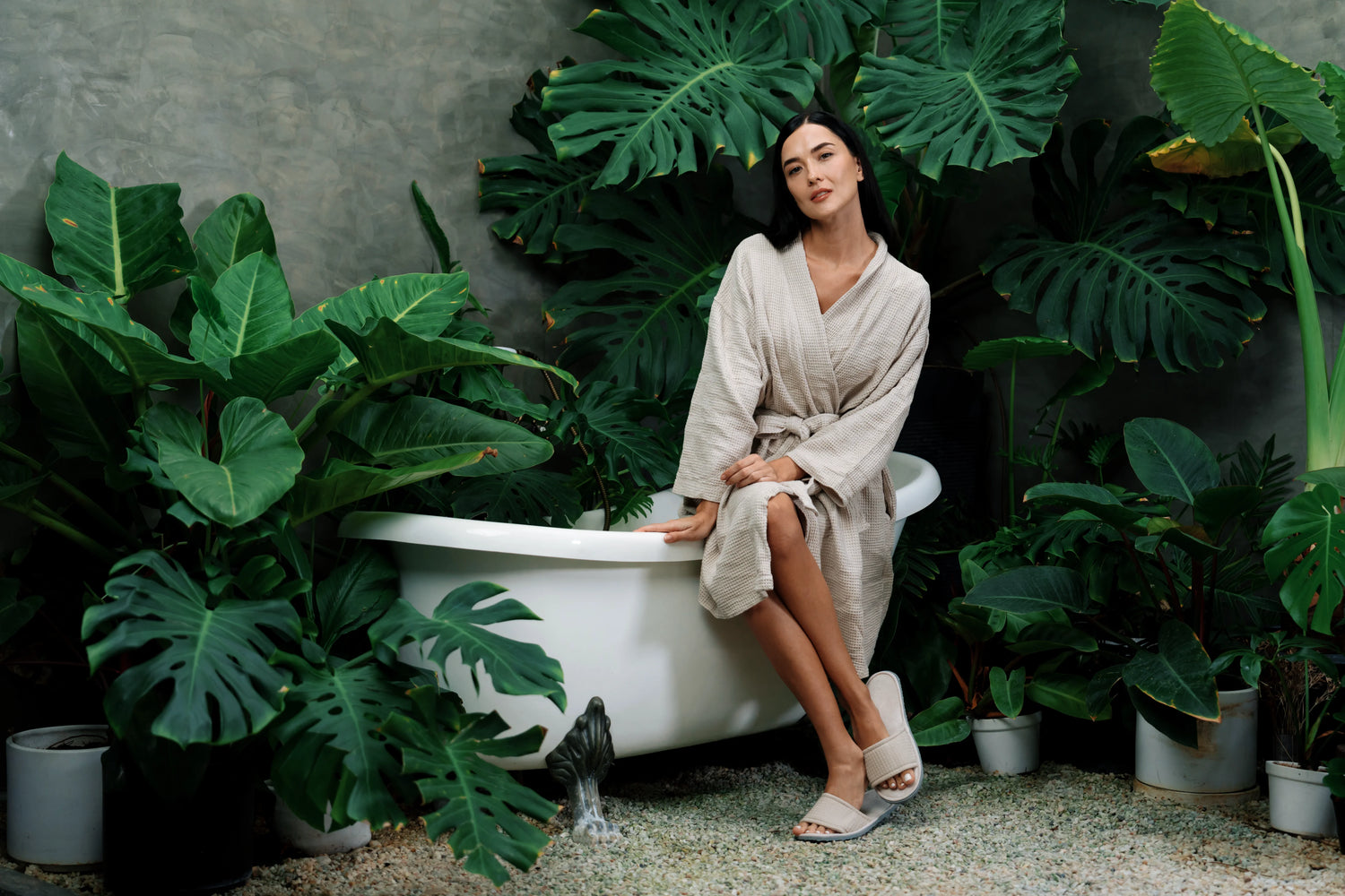 Woman in a bathrobe sitting by a bathtub surrounded by large green leaves